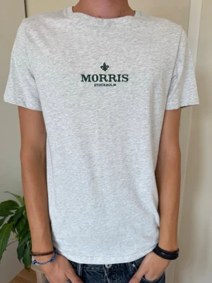 Ljusgrå t-shirt från Morris Stockholm - Snygg ljusgrå t-shirt från Morris Stockholm med diskret logga broderad i mörkgrönt på bröstet. Klassisk rund halsringning och korta ärmar. Tillverkad i mjuk bomull som känns skön mot huden. Perfekt till jeans eller shorts.