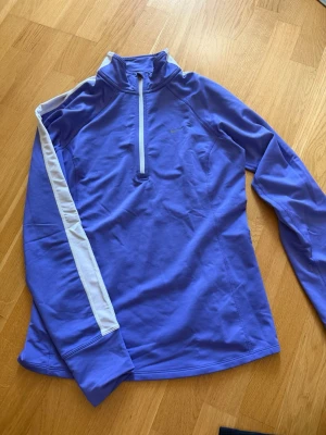 Lila Nike Dri-FIT träningströja - Snygg lila långärmad löpartröja från Nike med dragkedja framtill och vita detaljer längs ärmarna. Tillverkad i Dri-FIT-material som transporterar bort svett och håller dig torr under träningen. Perfekt för löpning eller annan träning. Har ett litet märke på framsidan vid bröstet, men syns knappt (se sista bilden). Annars mycket bra skick!