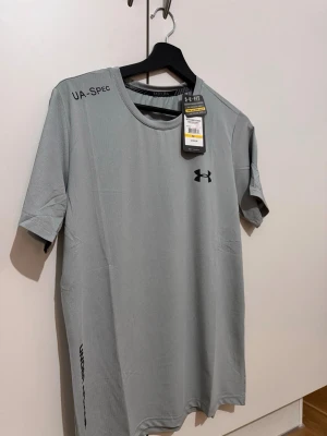 Grå träningströja från Under Armour - Snygg grå t-shirt från Under Armour med svart logga på bröstet och texten UA-SPEC på axeln. Tillverkad i ett lätt och snabbtorkande material, perfekt för träning. Rund halsringning och korta ärmar ger en sportig look.