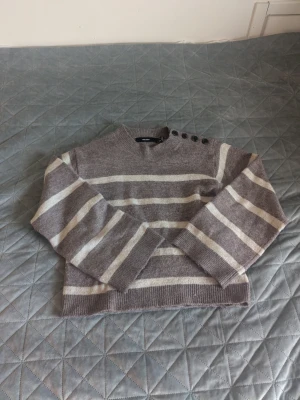 Beige/vit randig stickad tröja - Mysig stickad tröja i beige med vita horisontella ränder. Tröjan har rund halsringning och fyra dekorativa knappar på ena axeln. Långa ärmar och ribbade muddar vid ärmslut och nederkant. 