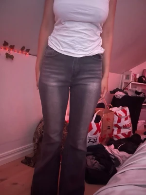 Grå bootcut jeans med detaljer - Säljer ett par grå bootcut jeans med snygga slitningar och broderade kors i rött och vitt med strass på bakfickorna. Jeansen har hög midja och klassisk femficksmodell. Perfekta för dig som vill sticka ut med coola detaljer.
