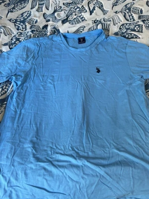 Ljusblå t-shirt från U.S. Polo Assn - Snygg ljusblå t-shirt från U.S. Polo Assn med klassisk passform och liten broderad logga på bröstet. Tillverkad i mjuk bomull och har rund halsringning samt korta ärmar. Perfekt för en avslappnad och stilren look.