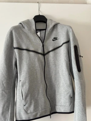 Grå Nike hoodie med dragkedja - Snygg grå hoodie från Nike med svart logga på bröstet och svarta detaljer. Tröjan har hel dragkedja, huva och fickor framtill samt en extra ficka med dragkedja på ärmen. Tillverkad i mjukt sweatshirtmaterial, perfekt för en sportig look.