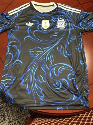 Argentina VM bortatröja - Snygg svart matchtröja med blått bladmönster och vita detaljer. Tröjan har vita ränder på axlarna, FIFA-märke och Argentinas landslagsemblem på bröstet. Tillverkad i ett lätt och ventilerande material, perfekt för fotboll eller träning.