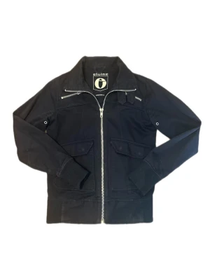 Cardigan från Elvine - Snygg mörkblå cardigan från Elvine med silverfärgad dragkedja, ribbade muddar vid ärmslut och midja samt två fickor med lock framtill. Jackan har en klassisk bomberkrage och detaljerade sömmar för en modern look.