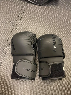 Svarta MMA-handskar från Chokem - Säljer ett par svarta MMA-handskar från Chokem i storlek L. Handskarna har vaddering över knogarna och bred kardborrestängning vid handleden för extra stöd. Perfekta för kampsportsträning och grappling.