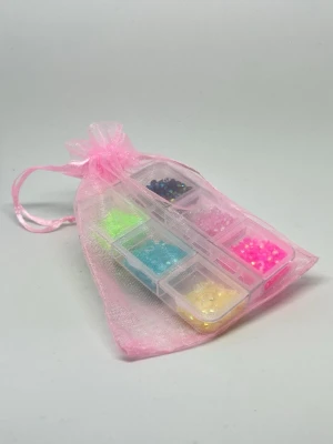 Mixade pärlor i rosa organzapåse - Säljer ett set med små, mixade pärlor i olika färger som neon, blå, gul, rosa och svart. Pärlorna kommer i en genomskinlig plastask med flera fack och levereras i en söt rosa organzapåse. Perfekt för att skapa egna smycken eller dekorera accessoarer.