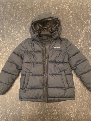 Svart pufferjacka från Everest - Snygg svart pufferjacka från Everest med huva och dragkedja framtill. Jackan har två stora fickor på sidorna och är vadderad för extra värme. Perfekt för kalla dagar och har en klassisk, stilren look.
