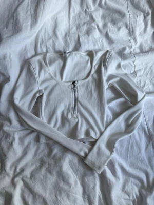 Vit croppad långärmad topp - Trendig vit croppad topp med lång ärm och rundad halsringning. Toppen har en ribbad struktur och en snygg dragkedja framtill som detalj. Perfekt att styla med höga jeans eller kjol för en cool look.