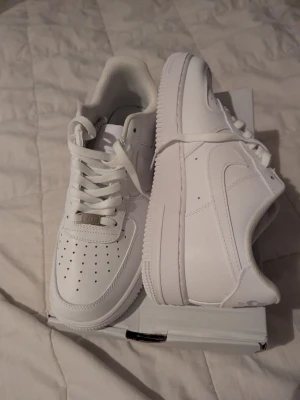 Nike Air Force 1 vita sneakers - Klassiska Nike Air Force 1 sneakers i helvitt läder med perforerad tå, vita skosnören och den ikoniska swoosh-loggan på sidan. Tjock sula med mönster för bra grepp och en stilren look som passar till det mesta.