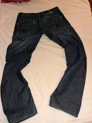 Mörkblå jeans herr längd 104 cm straight fit  - Säljer ett par mörkblå jeans med bra passform och slitna detaljer. Jeansen har två bakfickor med coola och  dekorativa sömmar och klassisk femficksdesign. Materialet är denim i bomull och byxorna har en avslappnad look. Dom är insydda. Dom är lite ljusare än bilderna ser ut/ konstigt ljus stora och bra fickor