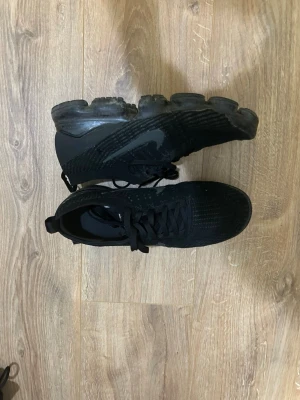 Svarta Nike VaporMax sneakers med luftig sula - Säljer ett par helsvarta Nike sneakers med stickad ovandel och genomskinlig, luftig sula. Skorna har klassisk snörning och diskret Nike-logga på sidan. Perfekta för dig som gillar sportig och stilren look.