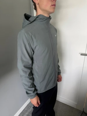 Grå vindjacka med huva från Nike - Snygg grå vindjacka från Nike med hel dragkedja och huva. Jackan har en minimalistisk design med Nike-logga på bröstet och är tillverkad i ett lätt, vindtätt material. Perfekt för blåsiga dagar och enkel att matcha med olika outfits.