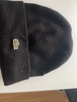 Svart mössa med broderad logga - Snygg svart mössa med uppvikt kant och en liten broderad krokodil-logga framtill. Klassisk och enkel design som passar till det mesta. Mössan är stickad och ser riktigt skön ut för kyliga dagar.