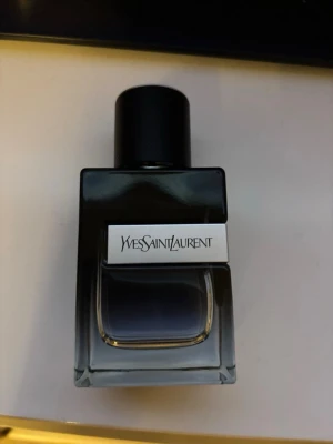 Yves Saint Laurent parfym herr - Säljer en stilren parfym från Yves Saint Laurent i en mörkgrå glasflaska med svart lock. Flaskan har en fyrkantig form och en metallisk etikett med loggan på framsidan. Perfekt för dig som gillar exklusiva dofter och snygg design.