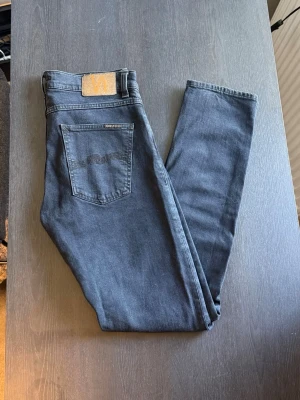 Mörkblå slim fit jeans från Nudie - Säljer ett par mörkblå slim fit jeans från Nudie Jeans med klassisk femficksdesign och diskret broderad bakficka. Jeansen har en smal passform och är tillverkade i mjukt denimtyg som sitter snyggt på benen. Perfekta för en clean och stilren look.