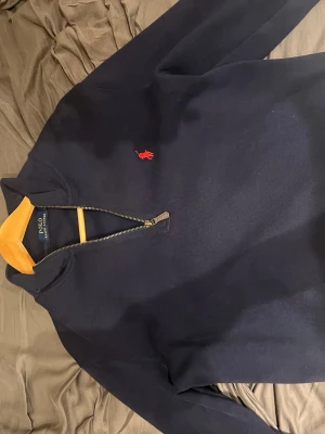 Mörkblå halvzip från Polo Ralph Lauren - Mörkblå tröja med halv dragkedja från Polo Ralph Lauren. Klassisk röd logga broderad på bröstet och gul insida på kragen. Tillverkad i mjukt material och har långa ärmar. Perfekt för en stilren och avslappnad look.