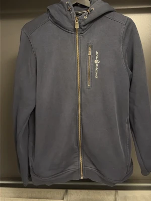 Marinblå hoodie från Sail Racing - Marinblå hoodie från Sail Racing med dragkedja framtill och bröstficka med zip. Snyggt tryck på bröstet och huva med snörning. Tillverkad i mjukt material, perfekt för chill dagar eller när du vill ha en clean look.