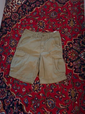 Vintage cargoshorts från Tommy Hilfiger - Snygga Vintage cargoshorts från Tommy Hilfiger med klassiska cargofickor på sidorna och diskret logga ovanför fickan. Tillverkade i mjuk bomull, perfekta för en avslappnad och trendig stil. Passar till allt från t-shirt till hoodie.