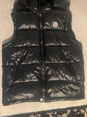 Svart dunväst från Moncler - Snygg svart dunväst från Moncler med glansig finish och två dragkedjefickor. Västen har hög krage och ikonisk Moncler-logga på bröstet. Perfekt för lager-på-lager och streetwear-stil.