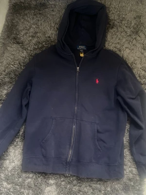 Mörkblå zip-hoodie från Polo Ralph Lauren - Mörkblå hoodie med dragkedja från Polo Ralph Lauren. Klassisk design med huva, kängurufickor och den ikoniska röda loggan broderad på bröstet. Tillverkad i mjukt bomullsmaterial som är skönt att ha på sig. Perfekt för en avslappnad och stilren look.