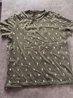 Olivgrön t-shirt från Polo Ralph Lauren - Säljer en olivgrön t-shirt från Polo Ralph Lauren med små vita broderade loggor över hela tröjan. Klassisk rund hals och korta ärmar. Tillverkad i mjuk bomull som känns skön mot huden. Perfekt för en avslappnad och stilren look. Knappt använd, skrynklig men de är bara o stryka den
