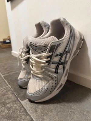Vita och grå Asics sneakers - Snygga vita och grå sneakers från Asics med mesh- och syntetmaterial. Skorna har klassisk låg profil, vita snören och svarta detaljer längs sidorna. Perfekta för en sportig och trendig look.