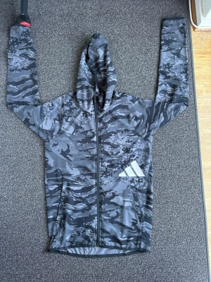 Grå camo hoodie med dragkedja - Snygg gråmelerad hoodie från Adidas med camomönster och hel dragkedja. Har huva och två fickor framtill samt Adidas-logga i vitt på ena sidan. Perfekt för en sportig och avslappnad stil.