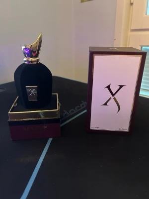 Lyxig parfymflaska med gulddetaljer - Säljer en exklusiv parfymflaska i svart med eleganta gulddetaljer och unik kork. Flaskan har en modern, stilren design och kommer i en matchande ask och förpackning med X-logga. Perfekt för dig som gillar lyxiga dofter och snygg inredning.