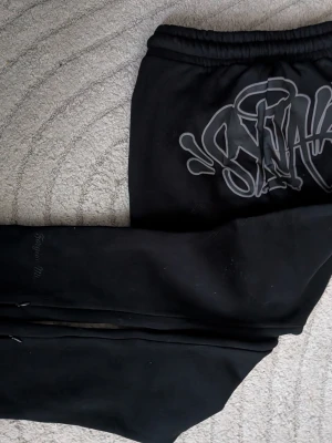 Black Sweatpants with Syna print - Svarta mjukisbyxor från LRG med stor grafisk print på ena benet. Tillverkade i mjukt, fleecefodrat material som känns skönt mot huden. Perfekta för en avslappnad streetwear-look.