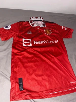 Manchester United matchtröja Ronaldo - Säljer en röd Manchester United matchtröja från Adidas med TeamViewer-logga på bröstet och Ronaldo 7 på ryggen. Tröjan har vita och svarta detaljer vid kragen och klubbmärke på bröstet. Perfekt för fotbollsfans! Detta är barn size 