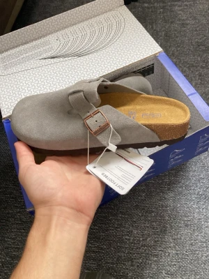 Grå mockatofflor från Birkenstock - Säljer ett par gråa Birkenstock tofflor i mocka med klassisk korksula och justerbart spänne i metall. Modellen har öppen häl och mjuk innersula för extra komfort. Perfekta för dig som gillar stilrena och bekväma skor. Helt nya oanvända , skickas samma dag 