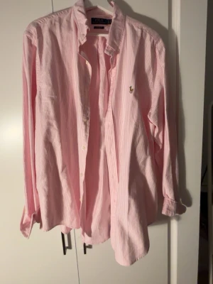  Polo Ralph Lauren skjorta - Säljer en rosa och vit randig skjorta från Polo Ralph Lauren i modellen Custom Fit. Skjortan har klassisk krage, knappar framtill och den ikoniska broderade loggan på bröstet. Perfekt för dig som gillar en stilren och tidlös look.