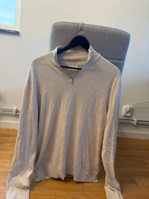 Beige stickad tröja med halv dragkedja - Beige stickad tröja med lång ärm och halv dragkedja vid halsen. Tröjan har en ribbad krage och muddar vid ärmslut och nederkant. Perfekt för lager-på-lager och enkel att matcha med olika outfits.