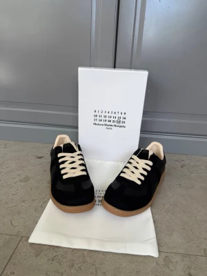 Maison Margiela svarta sneakers - Svarta sneakers från Maison Martin Margiela med beigea skosnören och innersula. Skorna har en klassisk låg siluett, ovandel i mocka och läder samt en gummisula i naturfärg. Perfekta för dig som gillar stilrena och tidlösa sneakers.