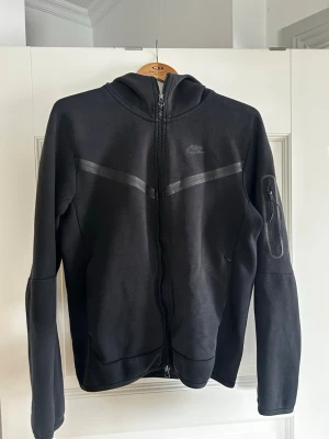 Svart Nike hoodie med dragkedja - Snygg svart hoodie från Nike med hel dragkedja framtill och huva. Har diskreta svarta detaljer över bröstet och en praktisk ficka med dragkedja på ena ärmen. Perfekt för en sportig och avslappnad stil. Pris kan diskuteras! 