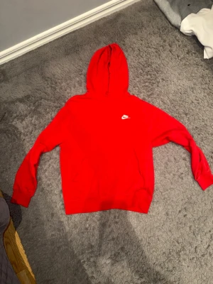 Röd Nike hoodie med huva - Snygg röd hoodie från Nike med klassisk vit logga på bröstet. Hoodien har huva med dragsko och en stor magficka. Perfekt för dig som vill ha en enkel men stilren look. Tillverkad i mjukt material för extra komfort.