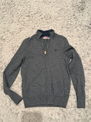 Morris Half-Zip - Mörkgrå stickad tröja med halv dragkedja och liten broderad logga på bröstet. Tröjan har ribbade muddar vid ärmslut och nederkant samt en hög krage. Perfekt för kyliga dagar och enkel att matcha med olika outfits. 100% Merino Ull.