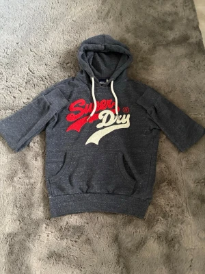Mörkgrå Superdry hoodie med kort ärm - Snygg mörkgrå hoodie från Superdry med korta ärmar och stor logga i rött och vitt framtill. Tröjan har huva med vita snören och en klassisk magficka. Perfekt för en avslappnad streetstyle-look.