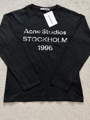 Svart långärmad topp Acne Studios - Svart långärmad topp från Acne Studios med tryck i vitt framtill där det står 'Acne Studios STOCKHOLM 1996'. Rund halsringning och mjukt material som känns skönt mot huden. Perfekt för en cool och avslappnad stil.