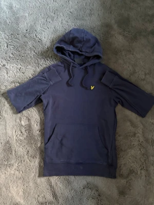 Mörkblå hoodie med korta ärmar - Snygg mörkblå hoodie med korta ärmar och huva. Har en gul broderad logga på bröstet och en stor magficka framtill. Tillverkad i mjukt bomullstyg, perfekt för en avslappnad streetwear-look.