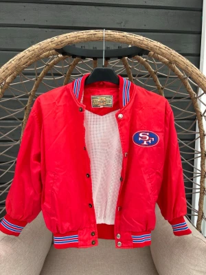 Retro bomberjacka strl. 128 - Röd bomberjacka med broderad SF-logga på bröstet. Jackan har blå och vita ränder på muddar och krage, tryckknappar framtill och ett sportigt meshfoder. Perfekt för dig som gillar retrostil.