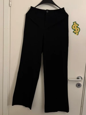 Svarta vida kostymbyxor - Säljer ett par svarta kostymbyxor med hög midja och vida ben. Byxorna har knapp och dragkedja framtill samt klassisk passform. Perfekta för en stilren och trendig look.