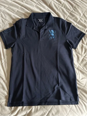 Marinblå pikétröja från Giordano - Snygg marinblå pikétröja från Giordano i slim fit-modell. Tröjan har krage, knappar framtill och ett blått broderat lejon på bröstet. Perfekt för en clean och sportig look. STORLEK XL ASIAN, PASSAR SOM M-L EUROPEISK STORLEK!!!