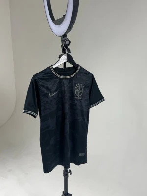 Svart Brasilien t-shirt Nike - Snygg svart t-shirt från Nike med Brasilien-logga och diskret mönster. T-shirten har korta ärmar, rund halsringning och vita detaljer på krage och ärmslut. Perfekt för dig som gillar sportig stil. Och är i super bra skick