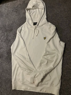 Vit hoodie från lyle & scout - Snygg vit hoodie med huva och dragsko. På bröstet sitter en liten gul logga i form av en fågel. Hoodien har känguruficka framtill och ribbade muddar vid ärmar och nederkant. Perfekt för en avslappnad och stilren look.