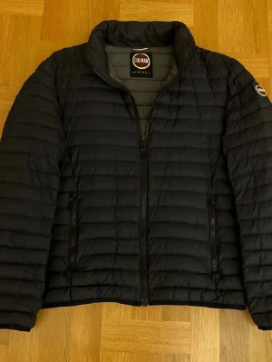 Svart dunjacka från Colmar - Snygg trendig svart dunjacka från Colmar med quiltad design och hög krage. Jackan har dragkedja framtill, två sidofickor med dragkedja och Colmar-logga på ärmen. Perfekt för kalla vinterdagar och riktigt stilren look. Storlek M men passar S