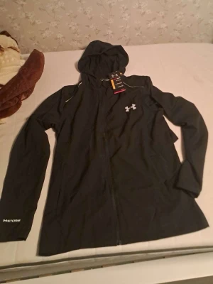 Svart träningsjacka med huva Under Armour - Svart träningsjacka från Under Armour med hel dragkedja och huva. Jackan har vita detaljer, logga på bröstet och texten 'STORM' på ärmen. Tillverkad i funktionsmaterial som passar perfekt för utomhusträning.
