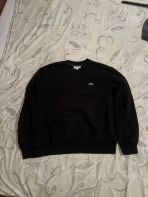Svart sweatshirt från Lacoste - Svart sweatshirt från Lacoste med klassisk rund halsringning och den ikoniska krokodilloggan broderad på bröstet. Tröjan har långa ärmar och ribbade muddar vid ärmslut och nederkant. Perfekt för en stilren och avslappnad look.
