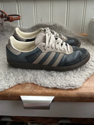 Adidas Samba skor, storlek 42 - Snygga sneakers i blått och grått med klassiska ränder på sidan. Skorna har mockadetaljer, vita snören och en mörk sula. Perfekta för dig som gillar retrostil och vill ha något som sticker ut lite extra. Använda ett flertal ggr med slitage på sulan men annars ok skick!